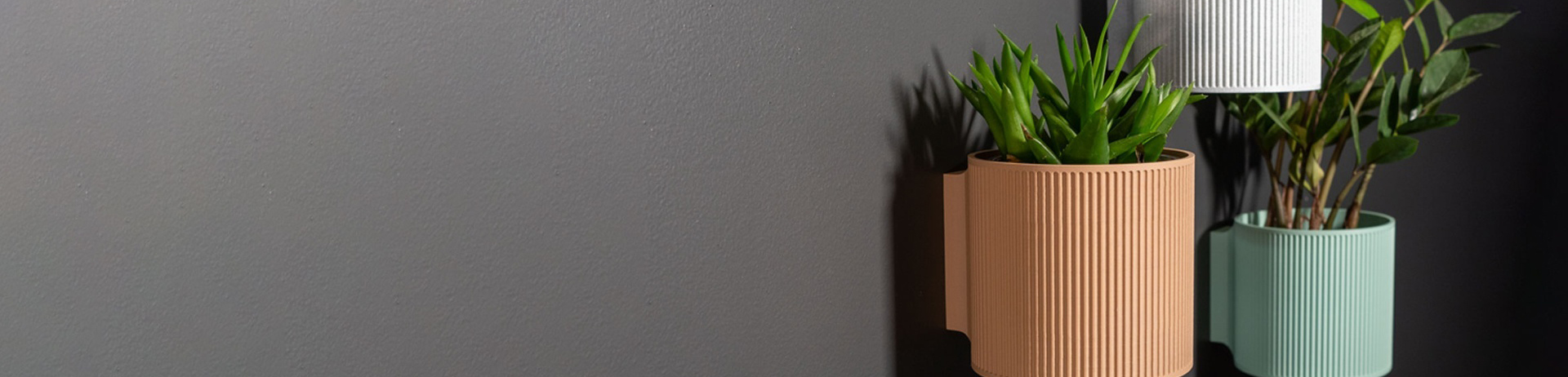 Tres macetas de pared impresas en 3D, en terracota, blanco y verde agua, montadas sobre una pared gris.